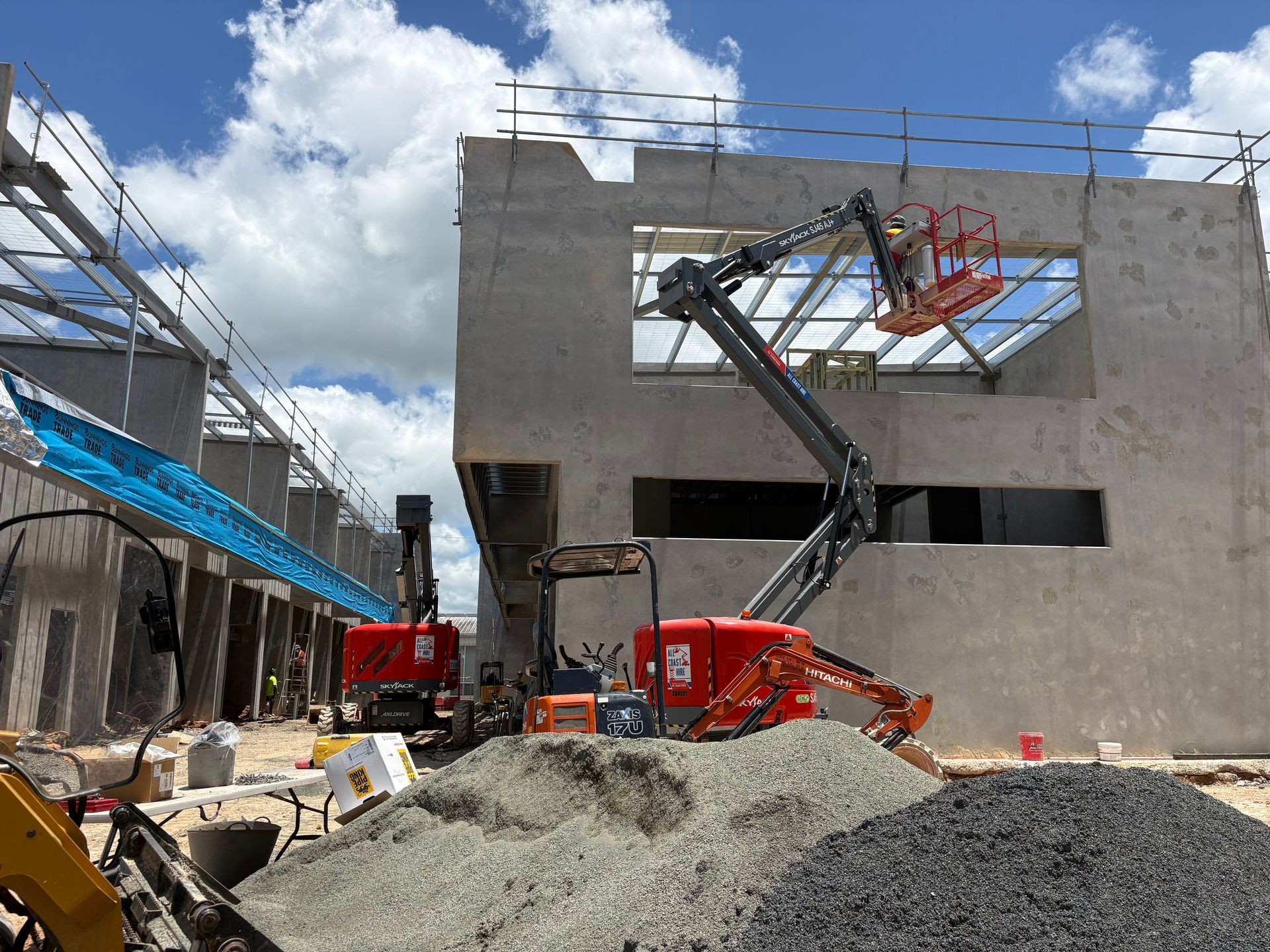 A person in a boom lift works on the exterior of a large, unfinished gray concrete building under a bright, cloudy sky.