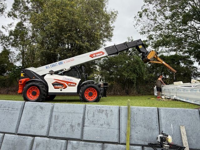 A white and orange Bobcat telehandler reaches toward a worker standing by construction materials on a grassy site.