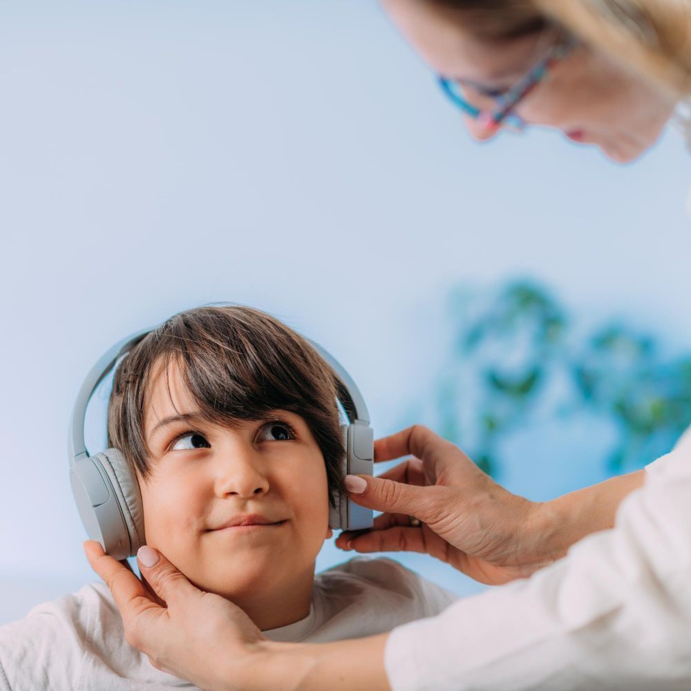Child wearing headphones while an adult adjusts them, with a soft blue background.