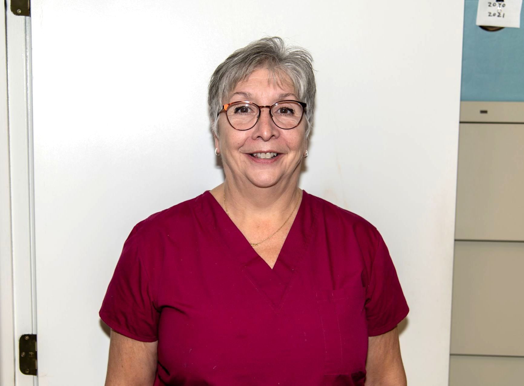 Smiling person in maroon scrubs standing indoors against a white wall