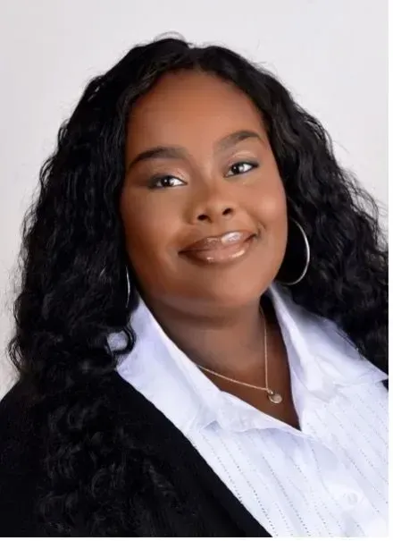 Smiling woman with long curly black hair in a white blouse and black jacket, posing against a light background