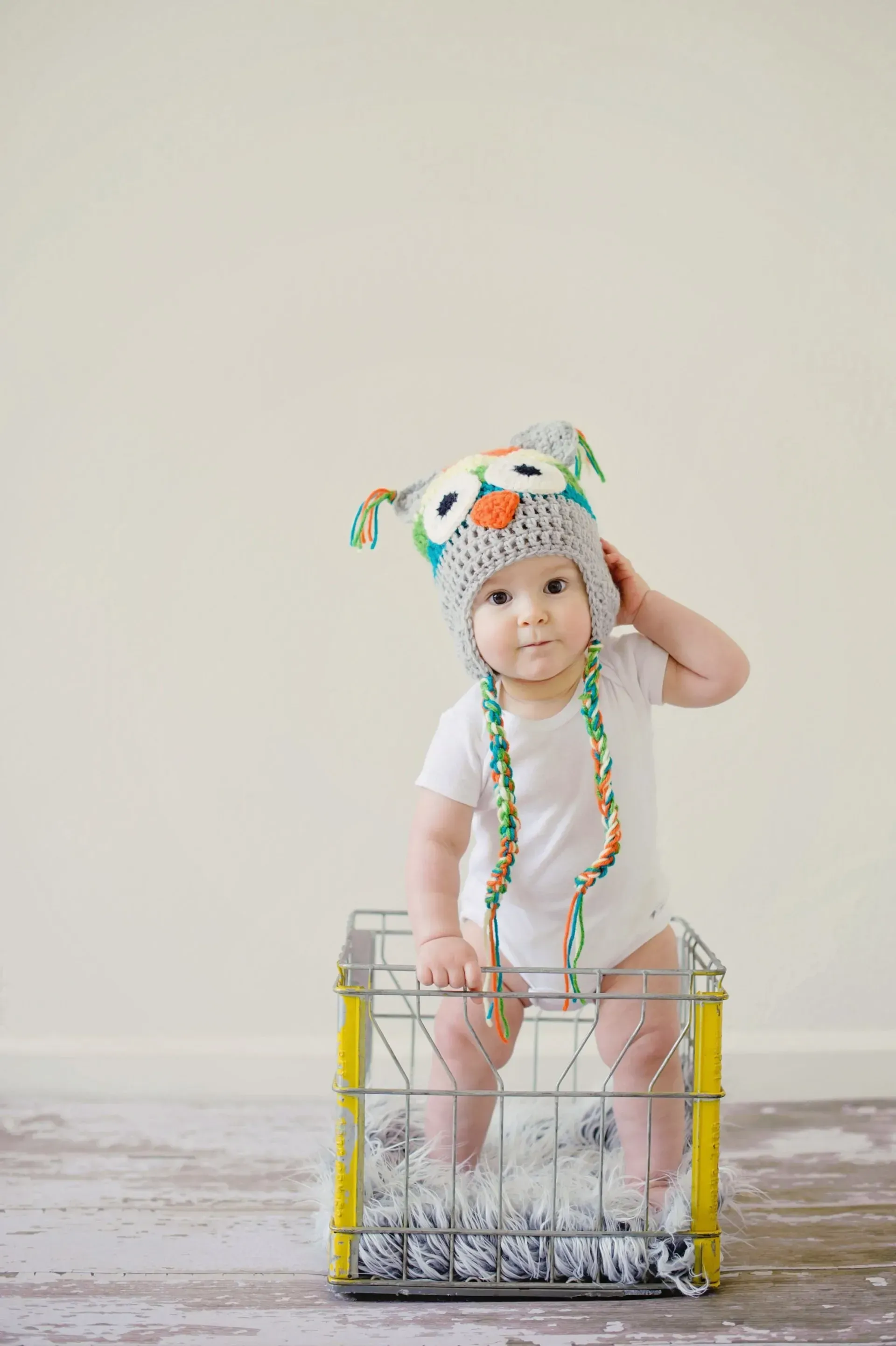 Baby in a colorful animal hat sitting in a clear crate against a white wall