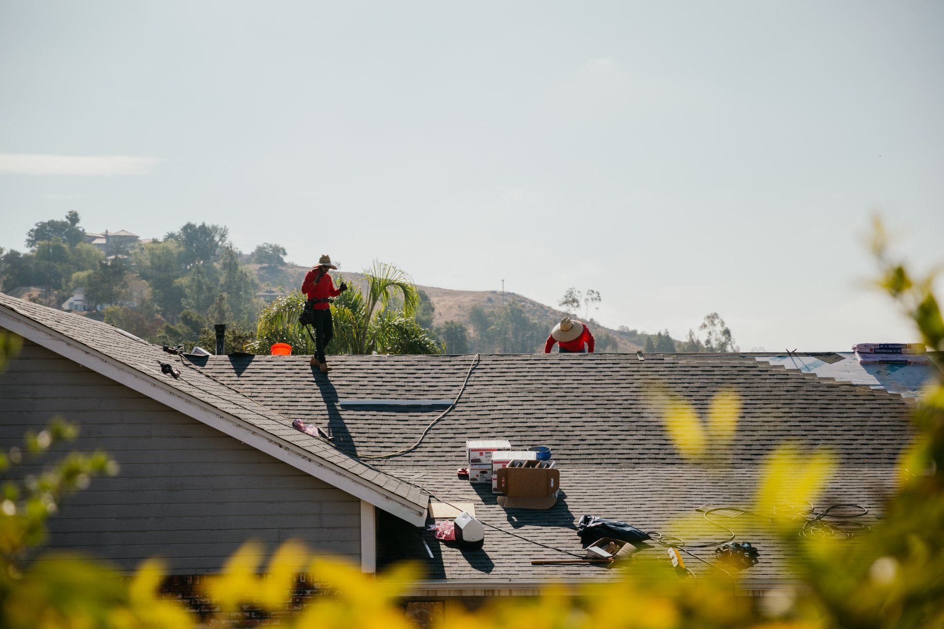 Roofers working on a house roof, with one in red near a ladder and another by a red bucket.