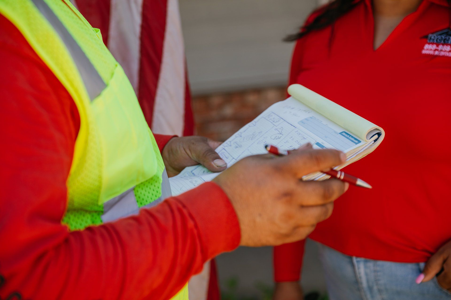 A person wearing a high-visibility vest writes on a notepad during a discussion with another person in a red shirt.