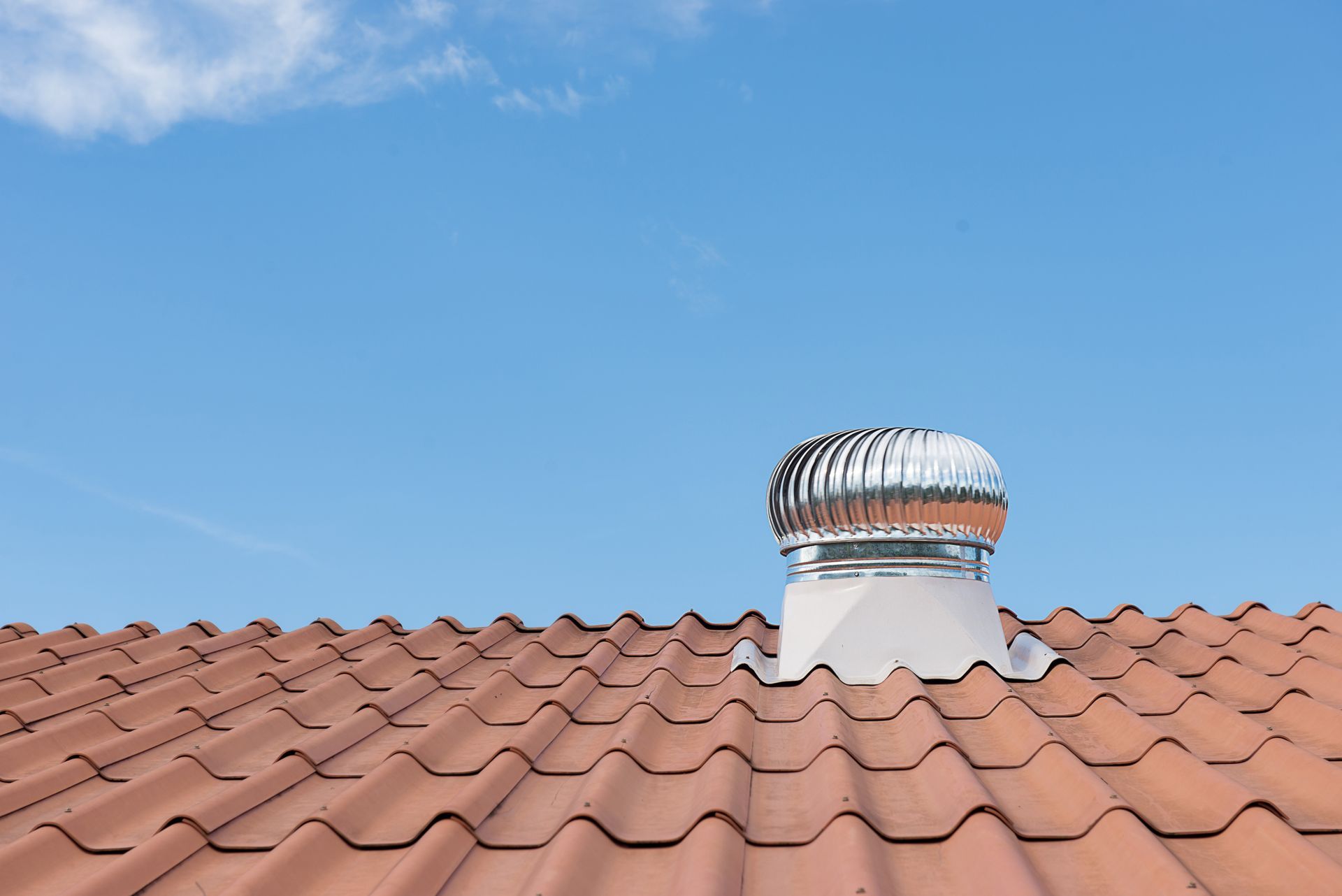 Rooftop vent with metal turbine cap above red clay roof tiles under a clear blue sky