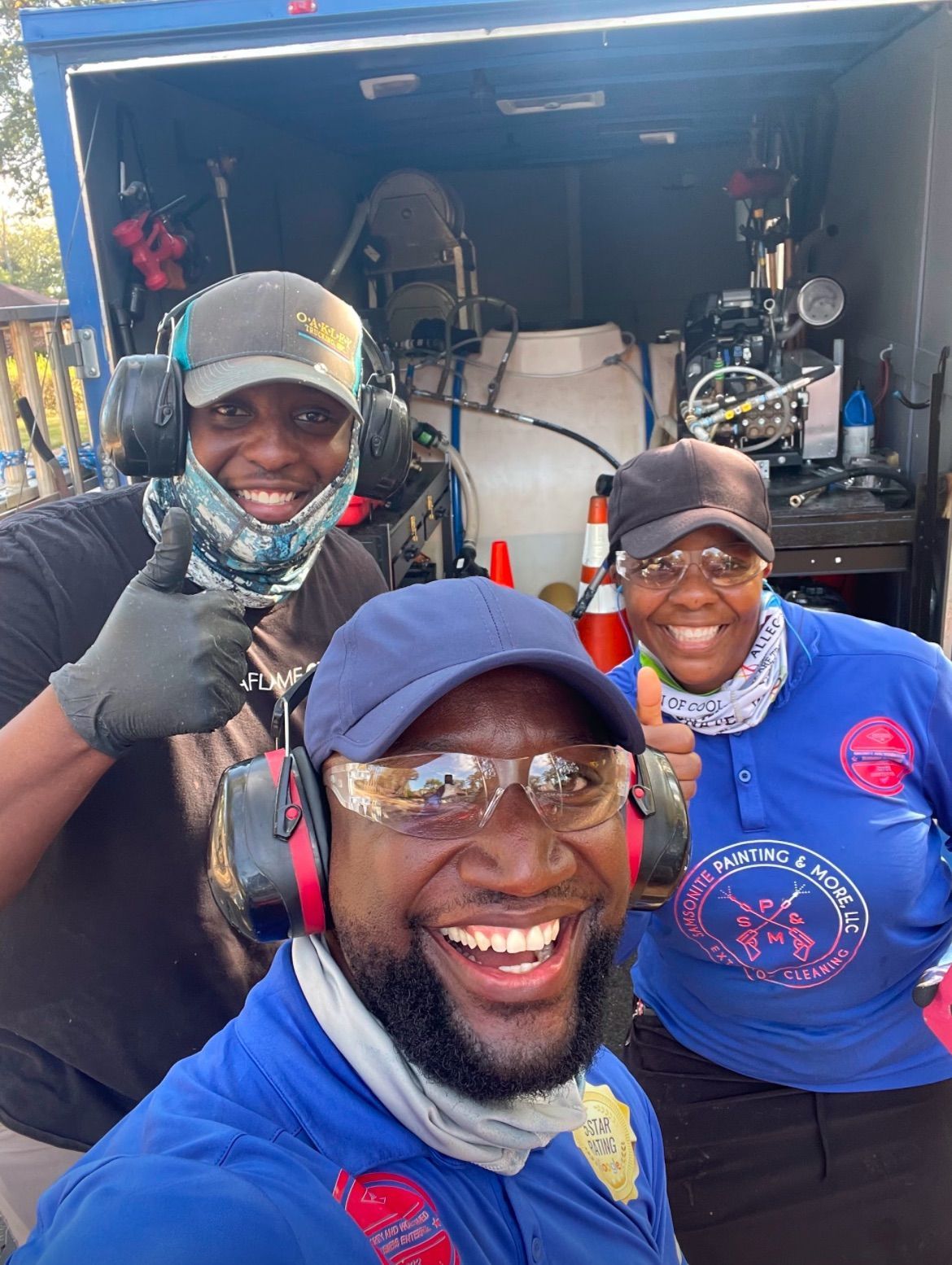 Three smiling people wearing safety gear giving thumbs up in front of a blue truck with equipment.