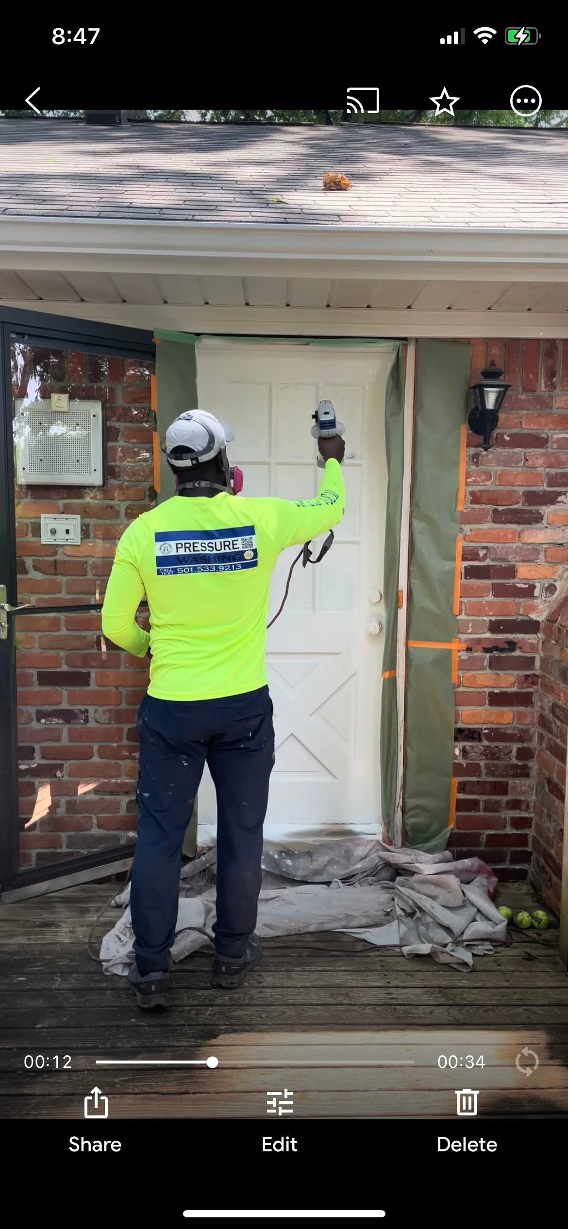 A worker wearing a neon green shirt paints a white door on a porch with brick siding.