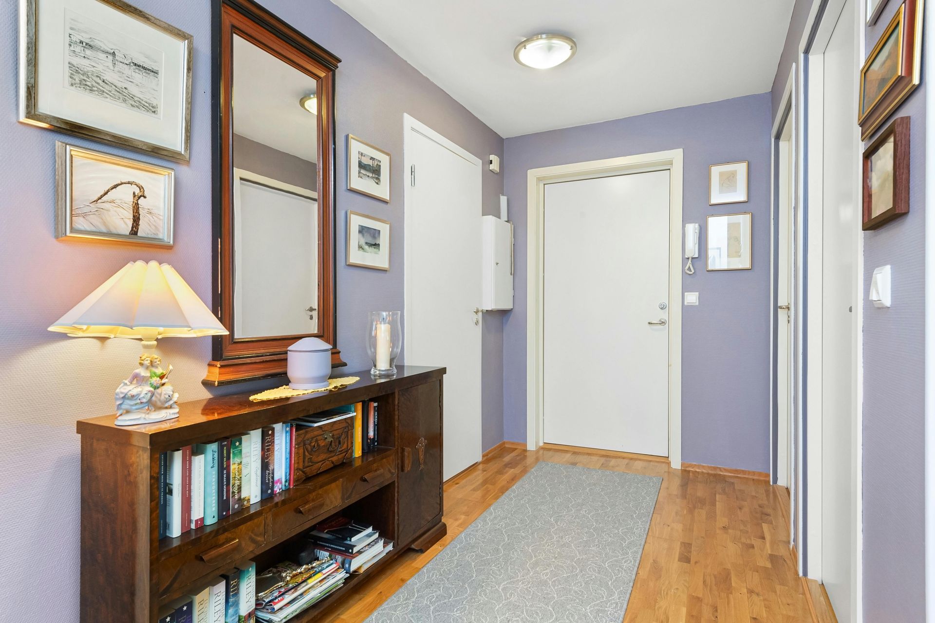 Hallway with wood floors and lavender walls. Features a dark wood cabinet with books, a mirror, and framed art.