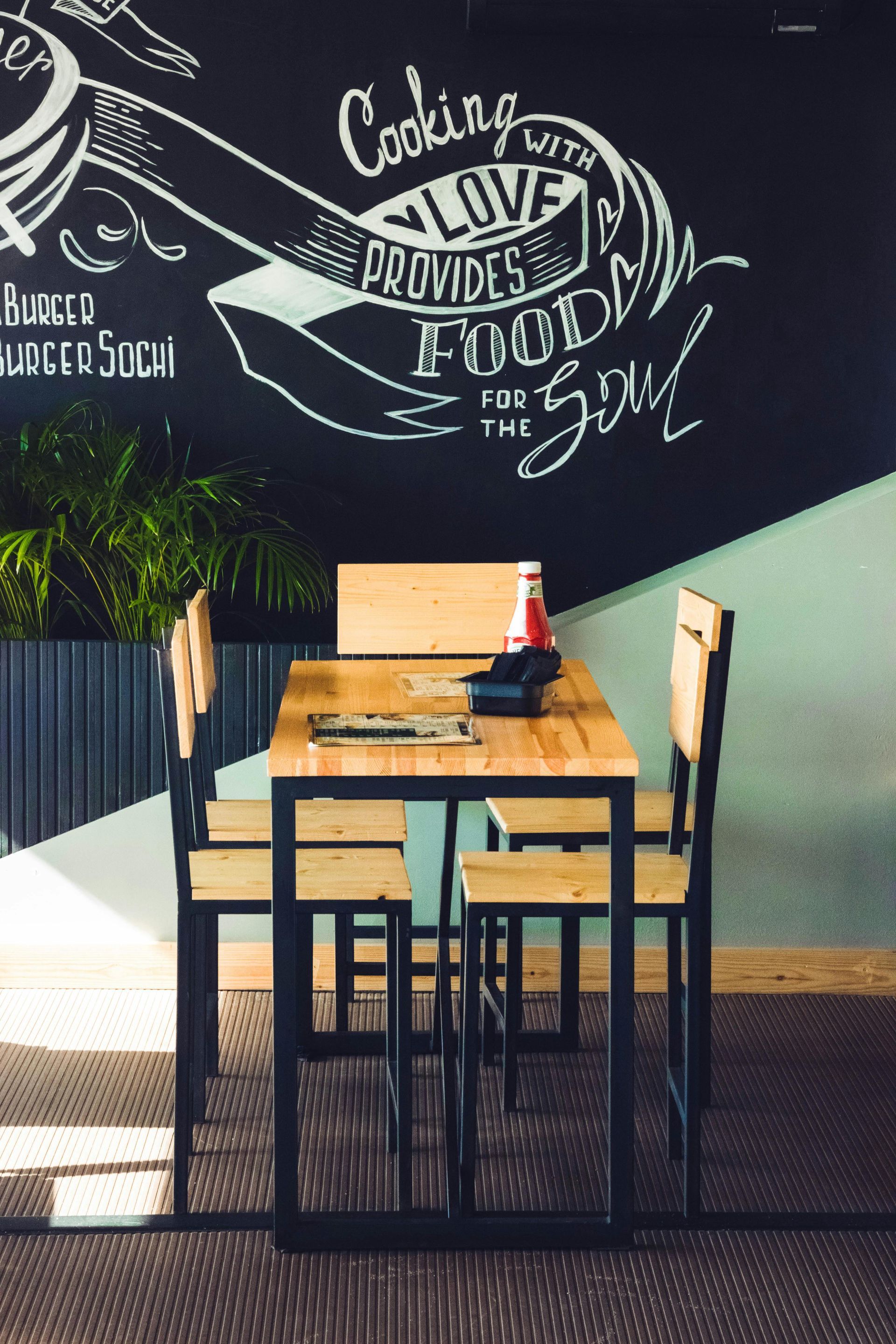 Restaurant dining area with a wooden table and chairs set against a chalkboard wall with food-related art and text.