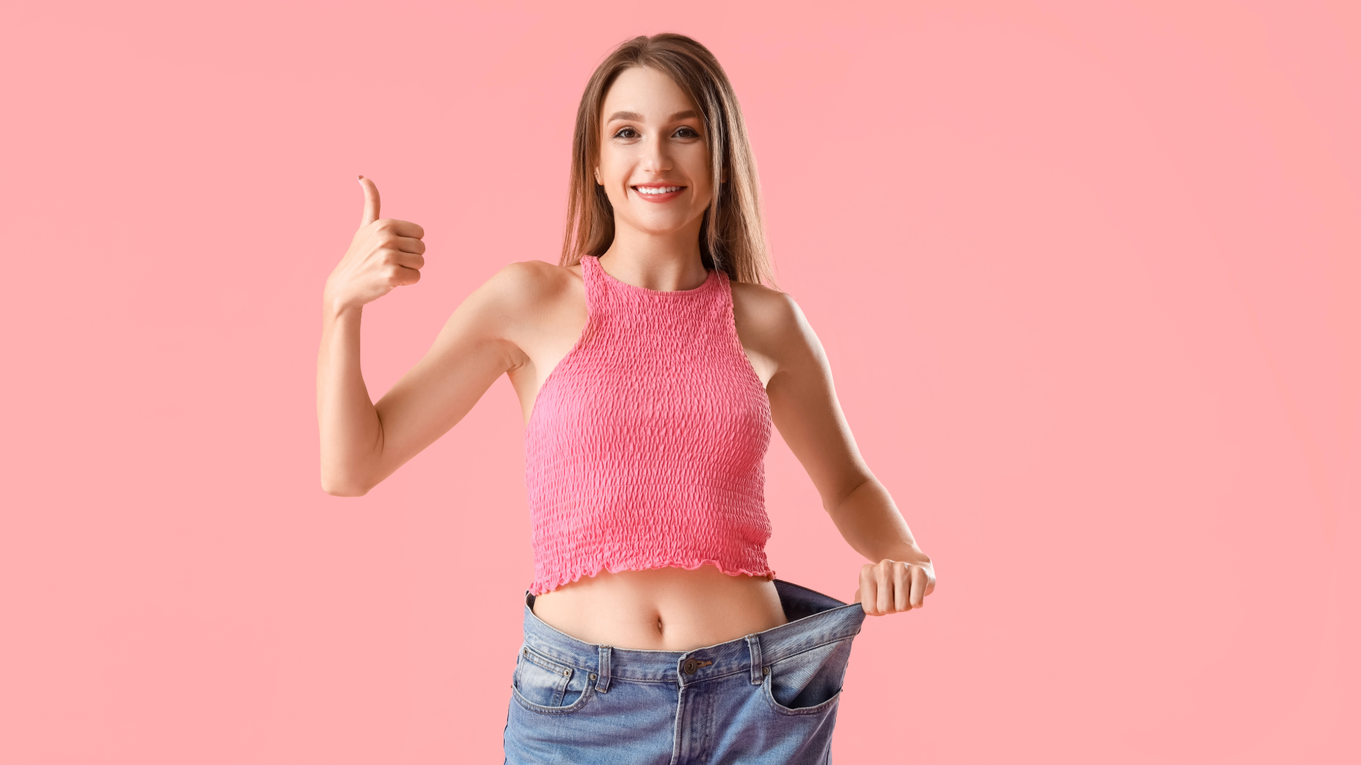 Woman in oversized jeans giving a thumbs-up, smiling, showing weight loss success against pink background.