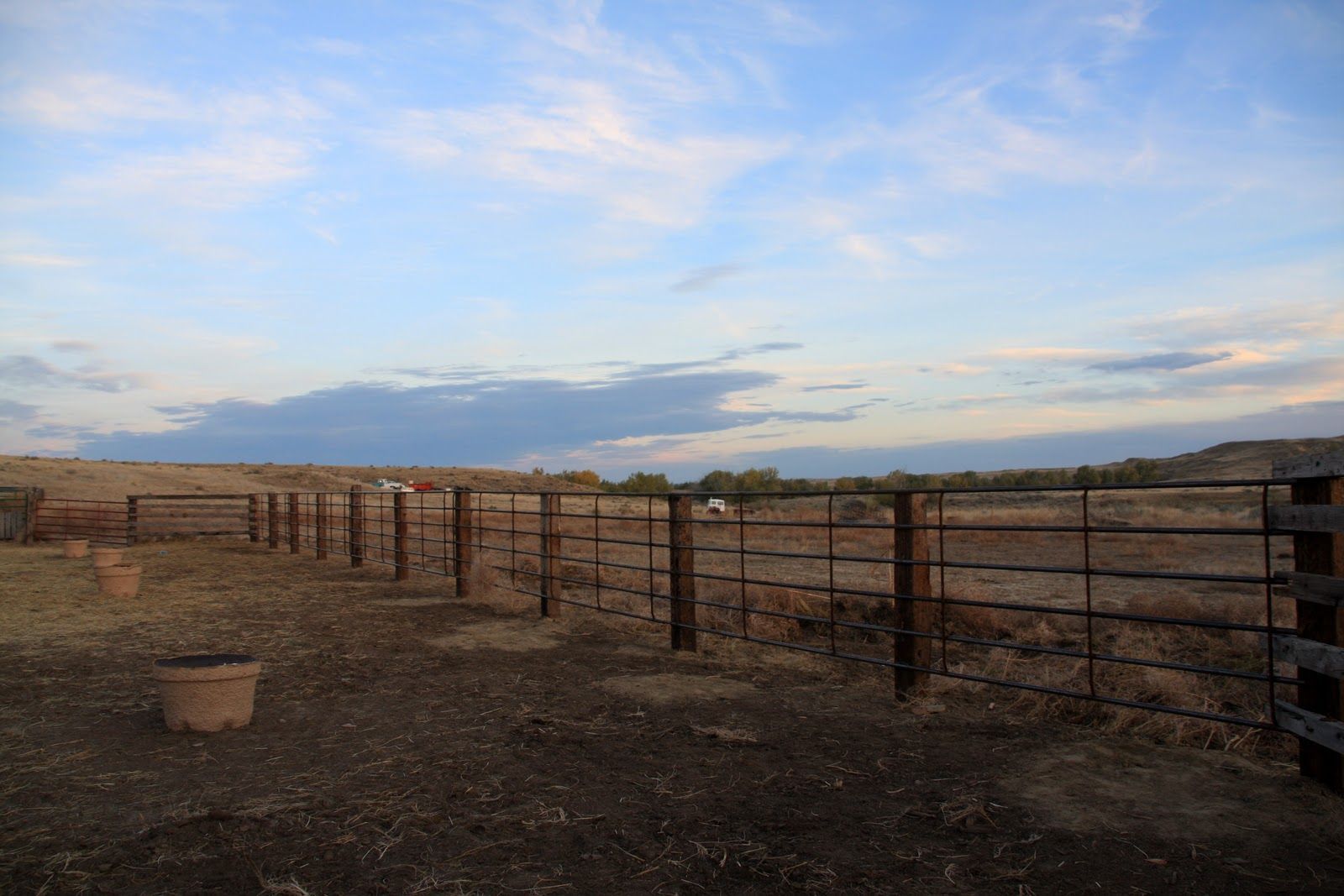 A rural landscape with a fence under a cloudy sky, brown earth, and empty plant pots.