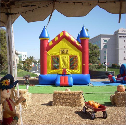 Bright inflatable bounce house at an outdoor fair, with hay bales and a small wagon of pumpkins in front.
