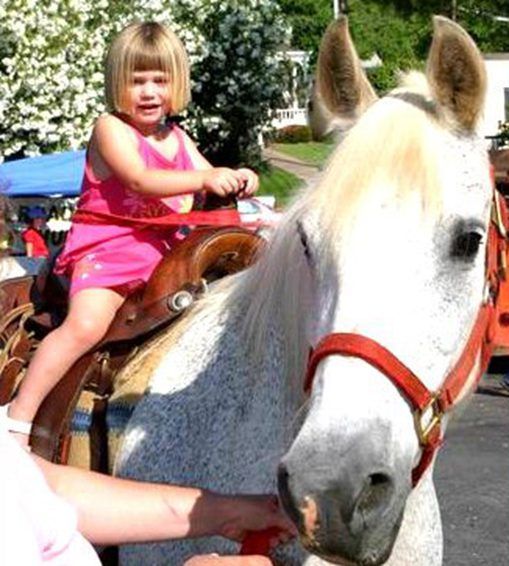 Toddler in pink dress riding a white horse with a red bridle outdoors