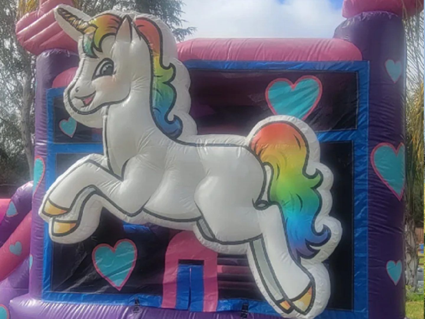 Unicorn bounce house with rainbow mane and tail, set against a dark background with pink hearts.