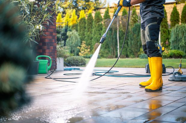 Person power washing a stone patio outdoors. Yellow boots, green foliage in the background.