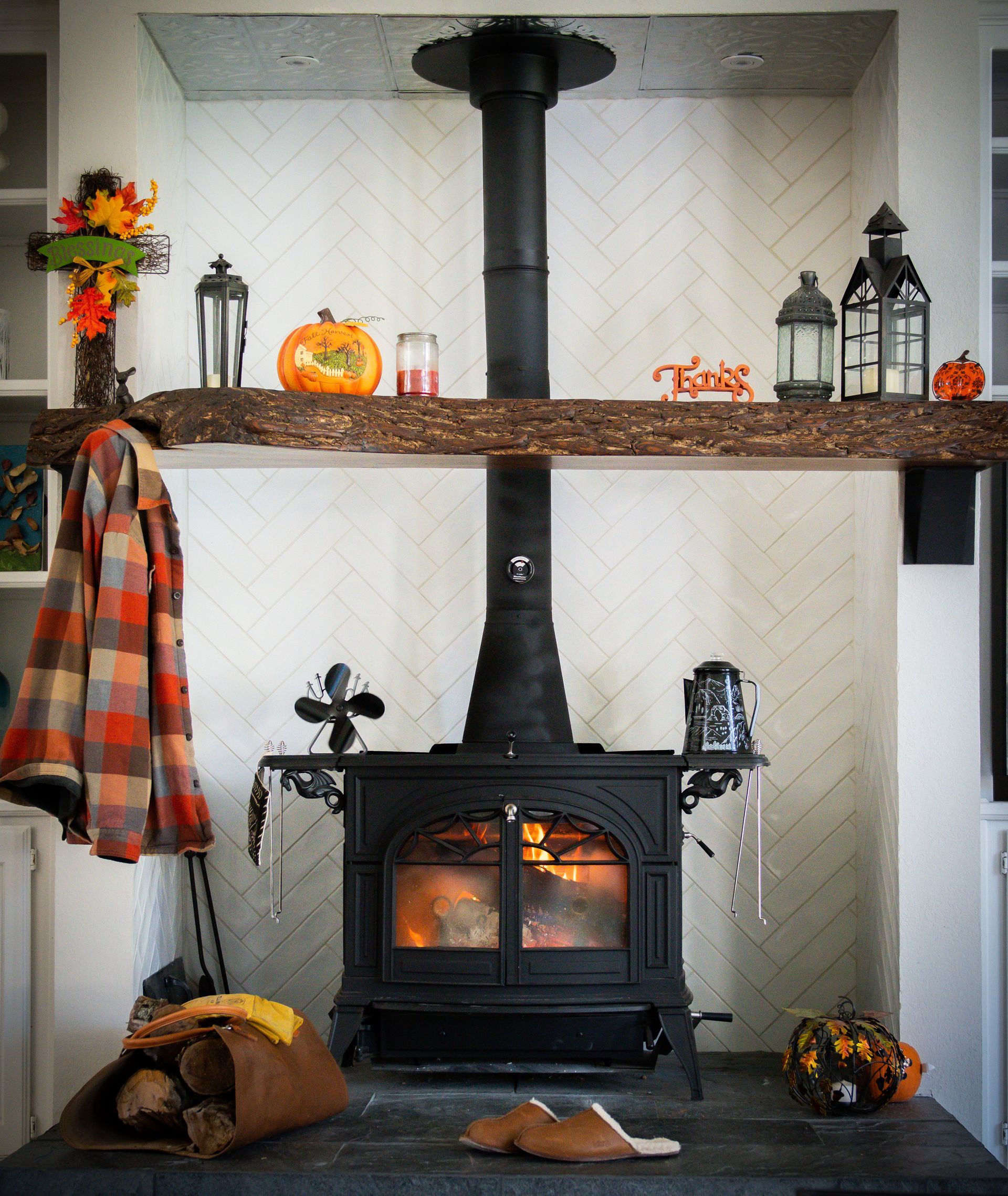 Cozy fireplace scene with black stove, wood mantel, and autumn decorations; a coat hangs nearby.