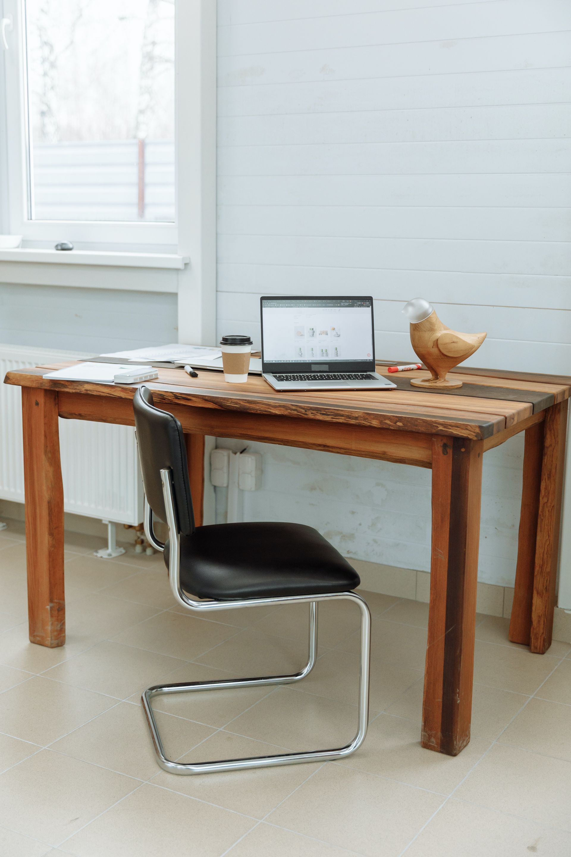 Wooden desk with a laptop, coffee, and a chicken statue, with a chair in front.