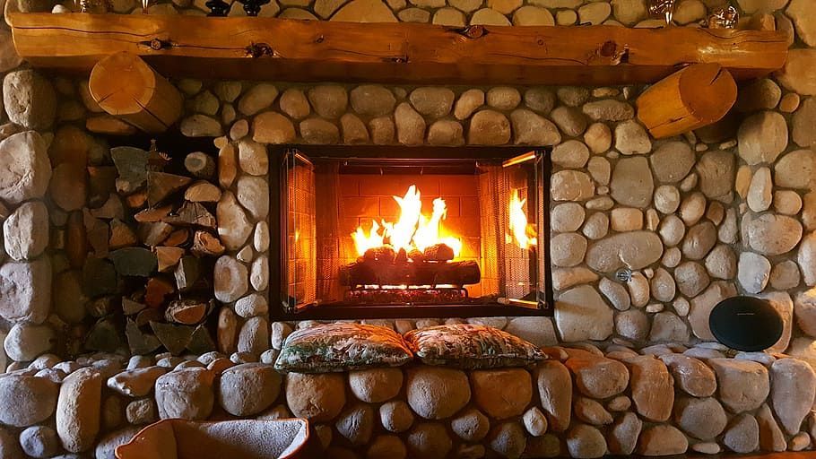 Fireplace with burning fire, surrounded by a stone facade, wooden mantle and firewood.