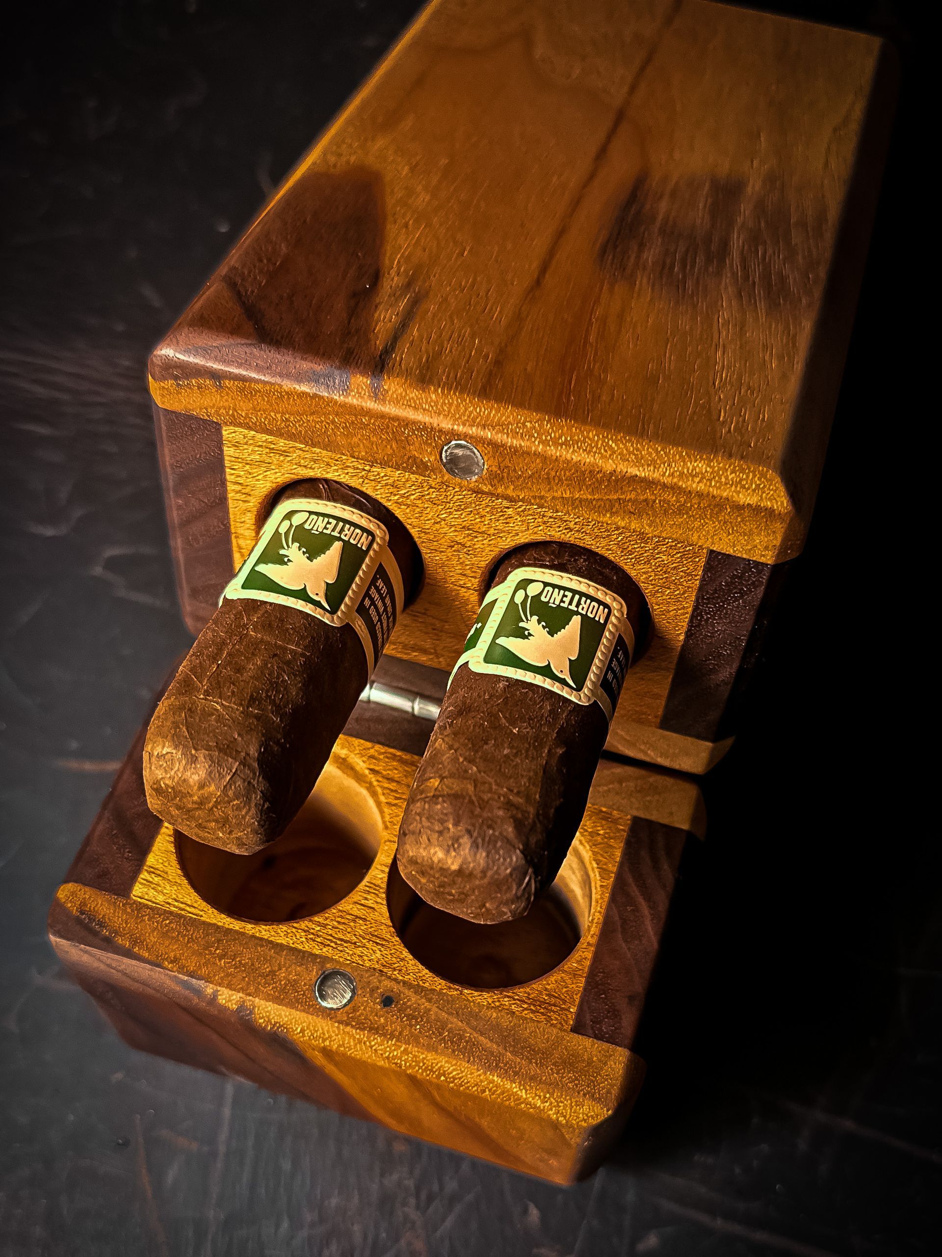 Two cigars in a wooden case. Brown with green labels, wood is dark and light brown, open.