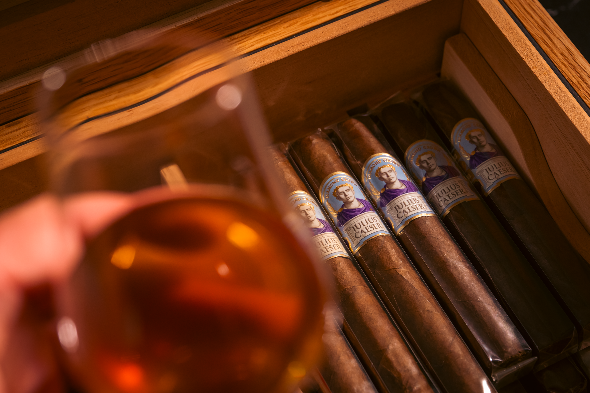 A glass of amber liquid in front of a humidor holding cigars with purple bands.