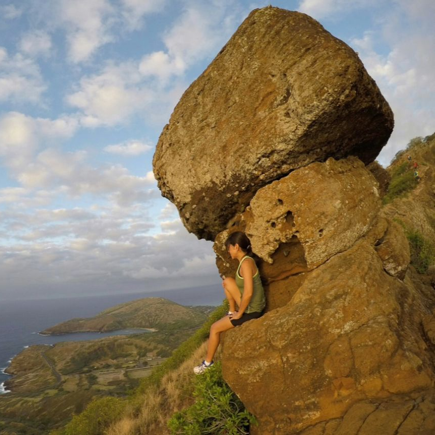 Woman sits on a large rock formation overlooking a coastal landscape.