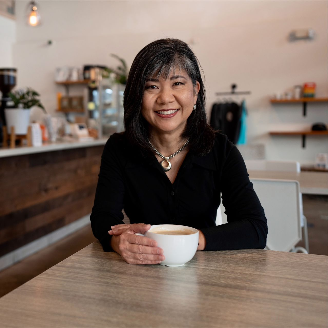 Woman holding a white mug smiling at the camera in a cafe setting with wooden table and counter.