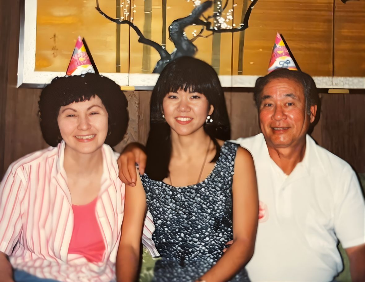 Three people wearing party hats, posing for a photo. A woman in a patterned dress is flanked by two older adults.