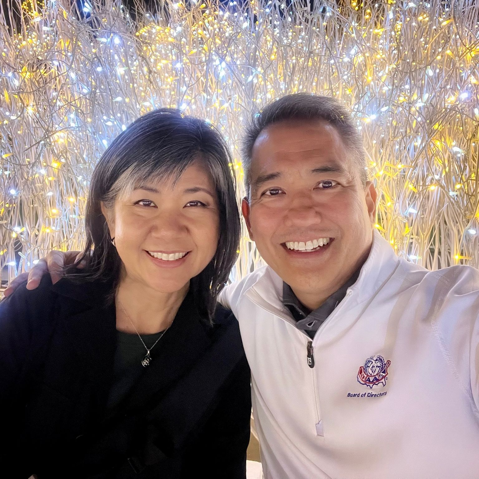 Couple smiling in front of a backdrop of white branches and glowing lights.