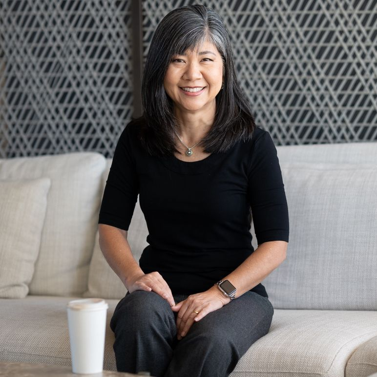 Woman smiling on a white couch, wearing a black top and gray pants. Coffee cup next to her.