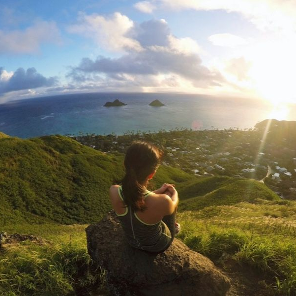 Woman sitting on rock, overlooking ocean and two small islands at sunset.