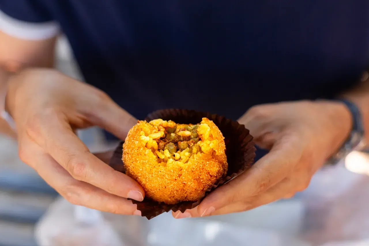 Arancini italiano aberto mostrando recheio de arroz, carne e ervilhas, com casca crocante dourada nas mãos de uma pessoa