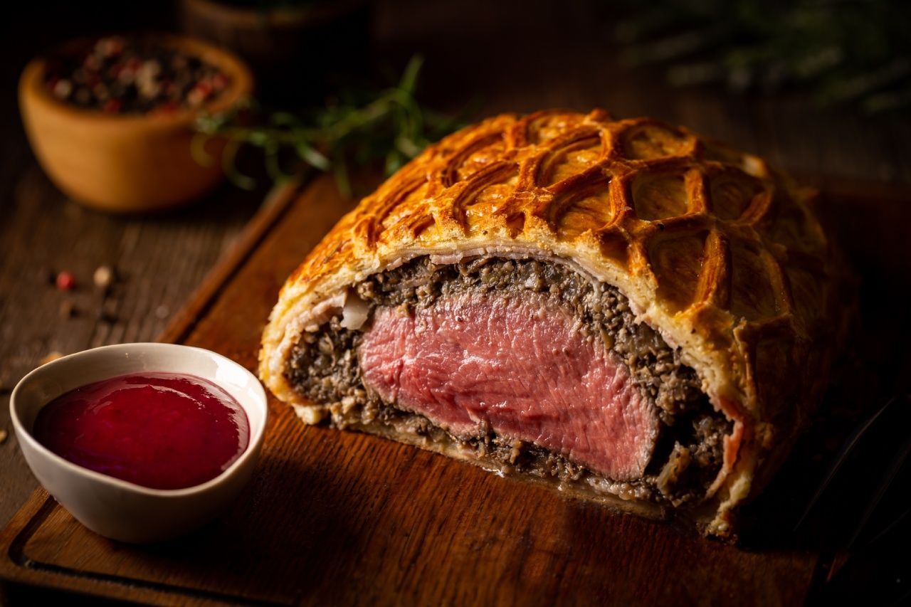 Um pedaço de carne está sobre uma tábua de corte de madeira ao lado de uma tigela de molho.