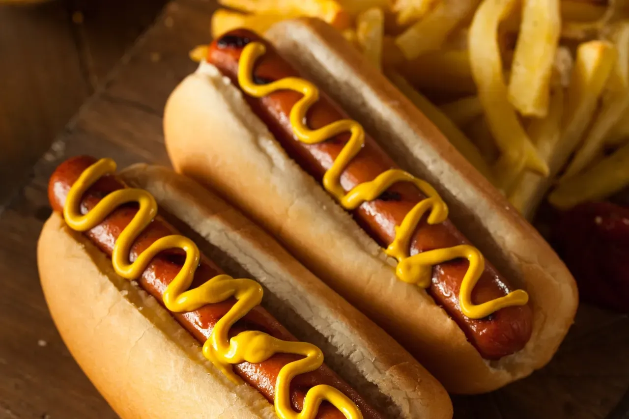 Cachorro-quente com salsicha grelhada no pão, coberto com mostarda, servido ao lado de batatas fritas.