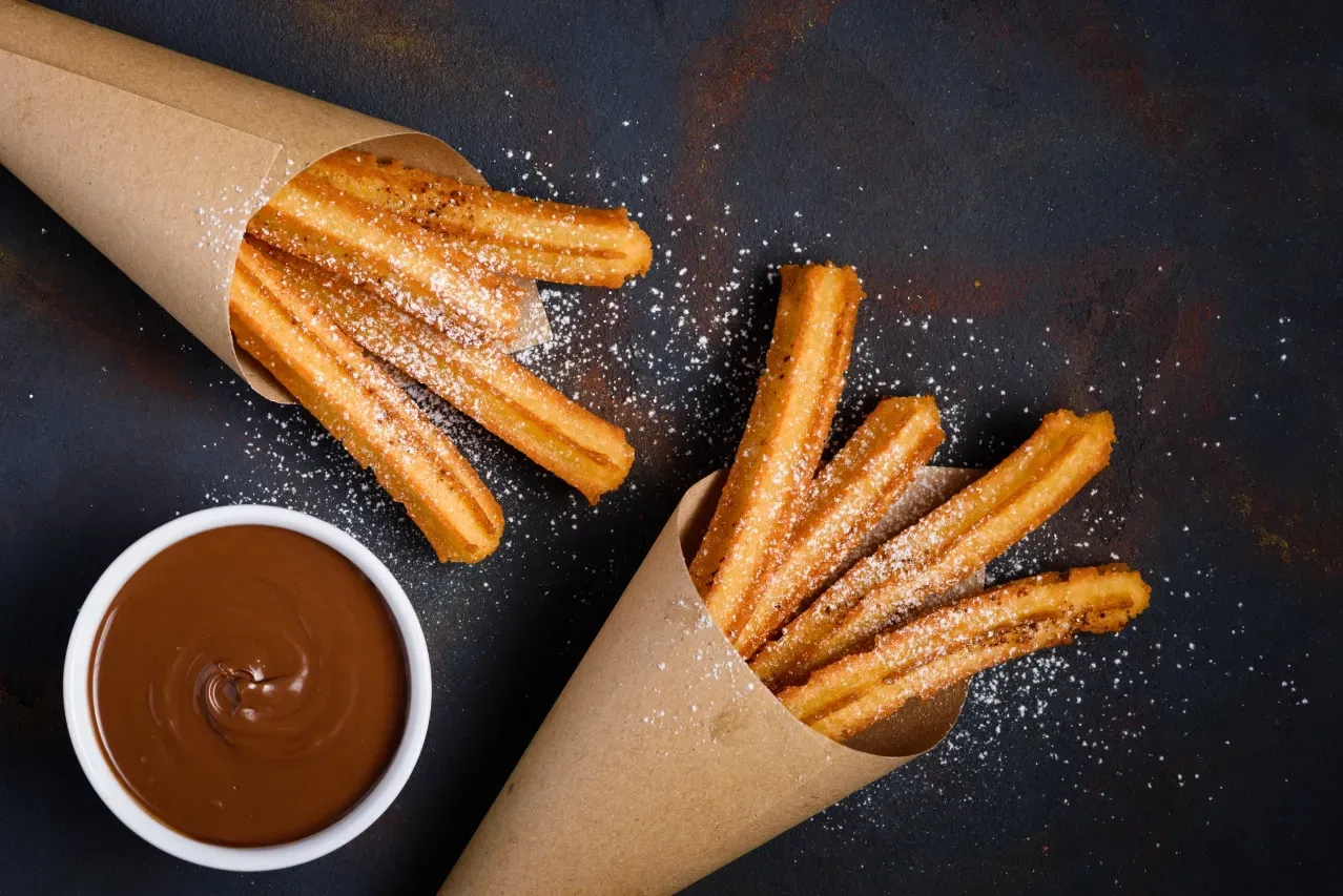 Churros em cones de papel ao lado de uma tigela de calda de chocolate.