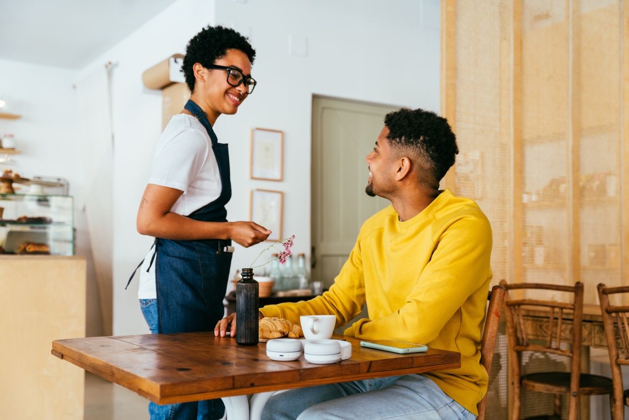 Atendente sorridente conversa com cliente em cafeteria enquanto serve café e croissant sobre a mesa.