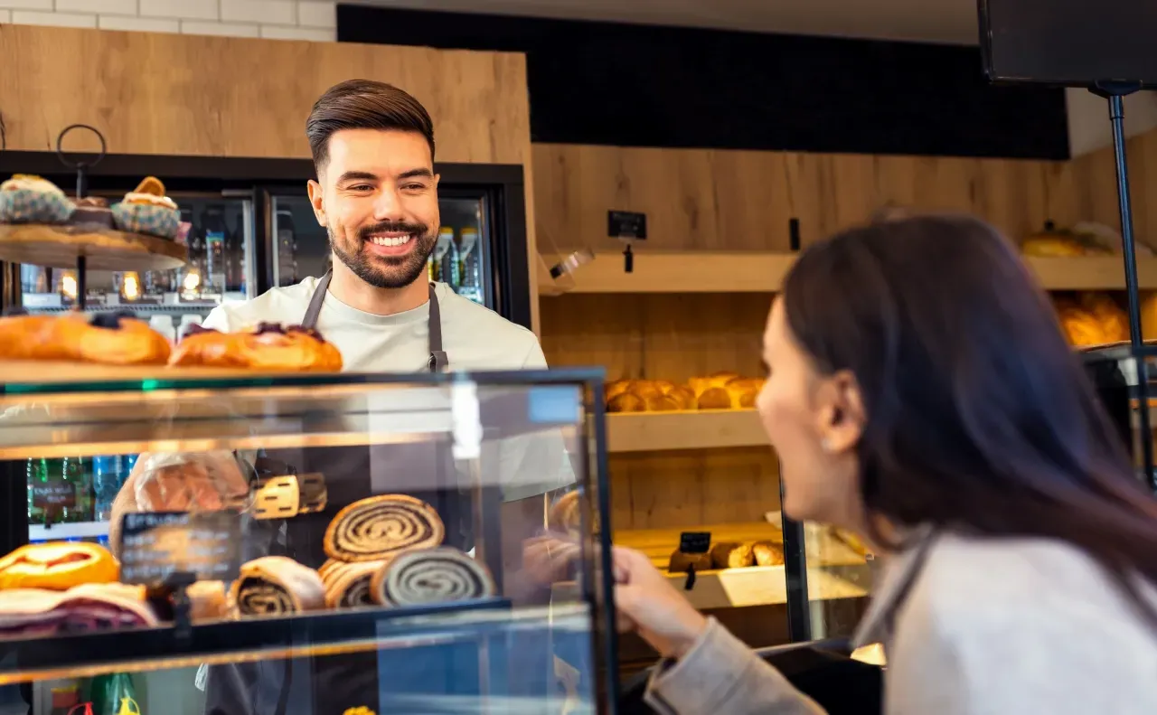 Atendimento em padaria com balconista sorrindo e cliente escolhendo pães frescos no balcão.