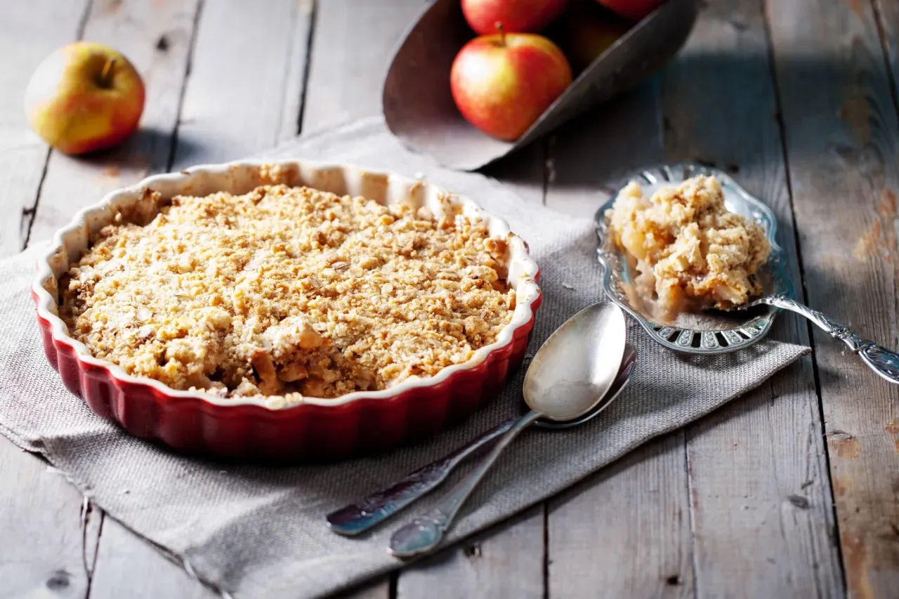 Crumble de maçã sobre mesa de madeira com maças e talheres