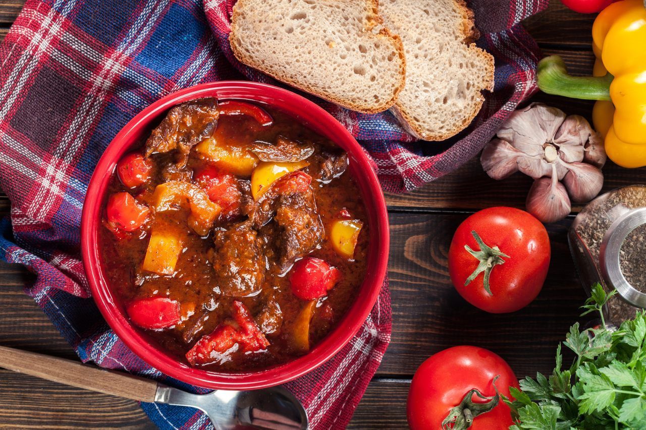 Uma tigela de ensopado de carne com vegetais e pão sobre uma mesa de madeira.