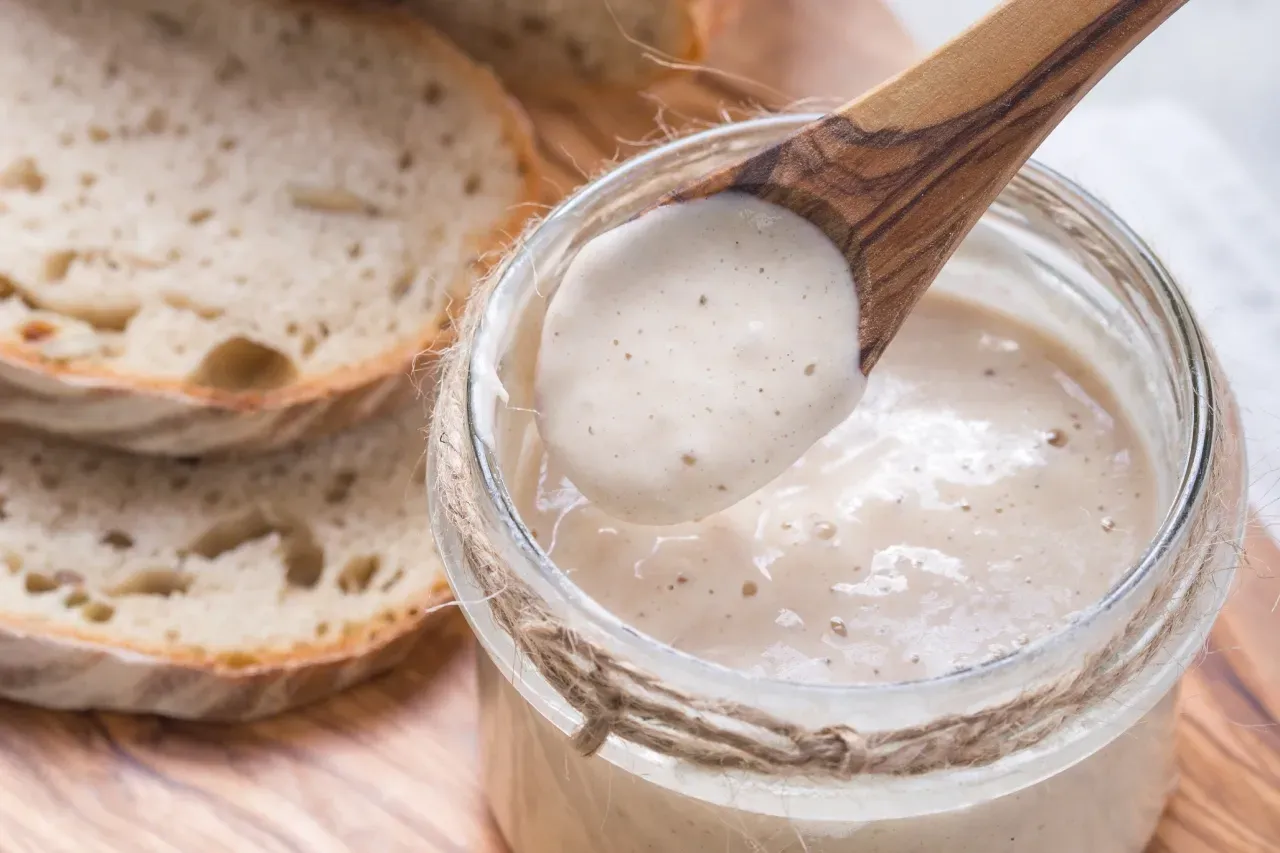 colher de pau em pote contendo fermento natural com fatias de pães artesanais, sobre tábua de madeir