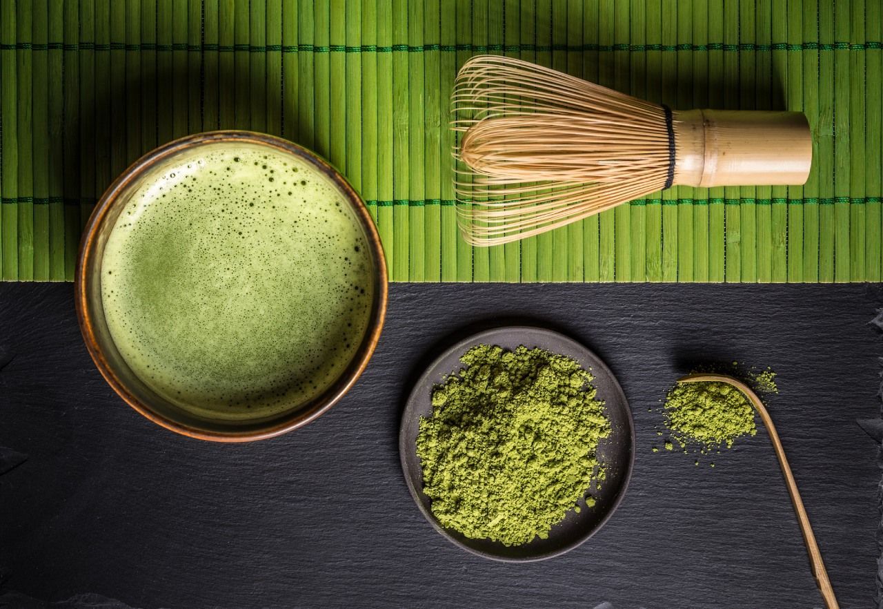 Uma tigela de chá verde, um batedor e uma colher de pó de chá verde sobre uma mesa.