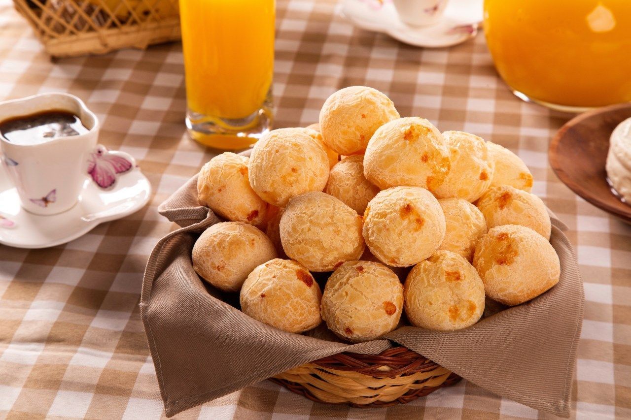 pães de queijo assados em cesto sobre mesa com toalha, xícara de café e copo de suco de laranja