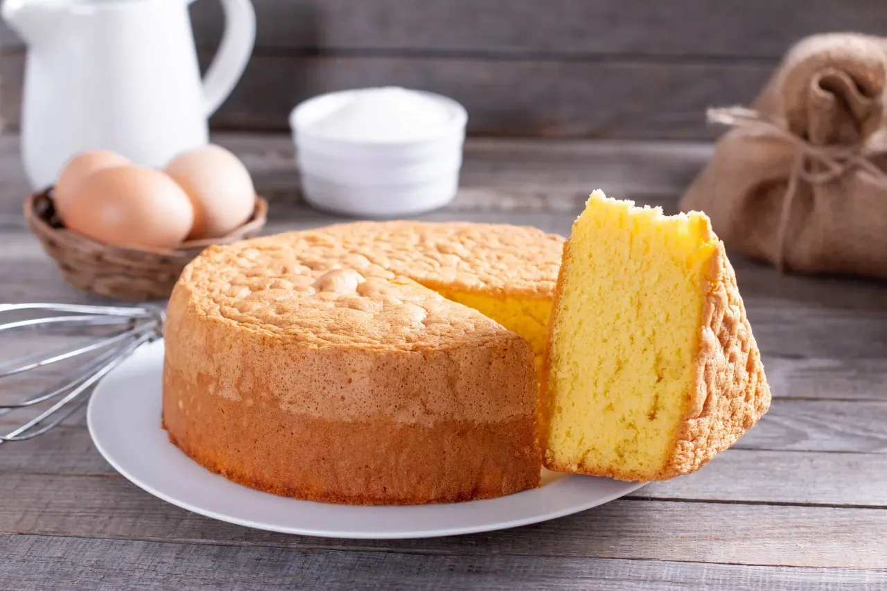 Pão de ló fofinho e alto sobre prato branco, com uma fatia cortada mostrando o miolo leve e aerado. 