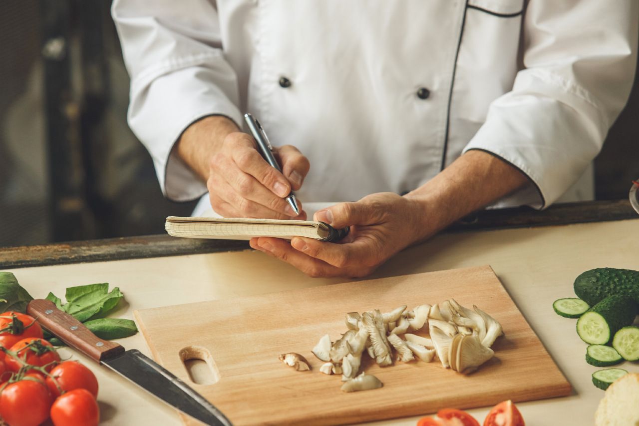 Um chef está escrevendo em um caderno enquanto corta vegetais em uma tábua de corte.
