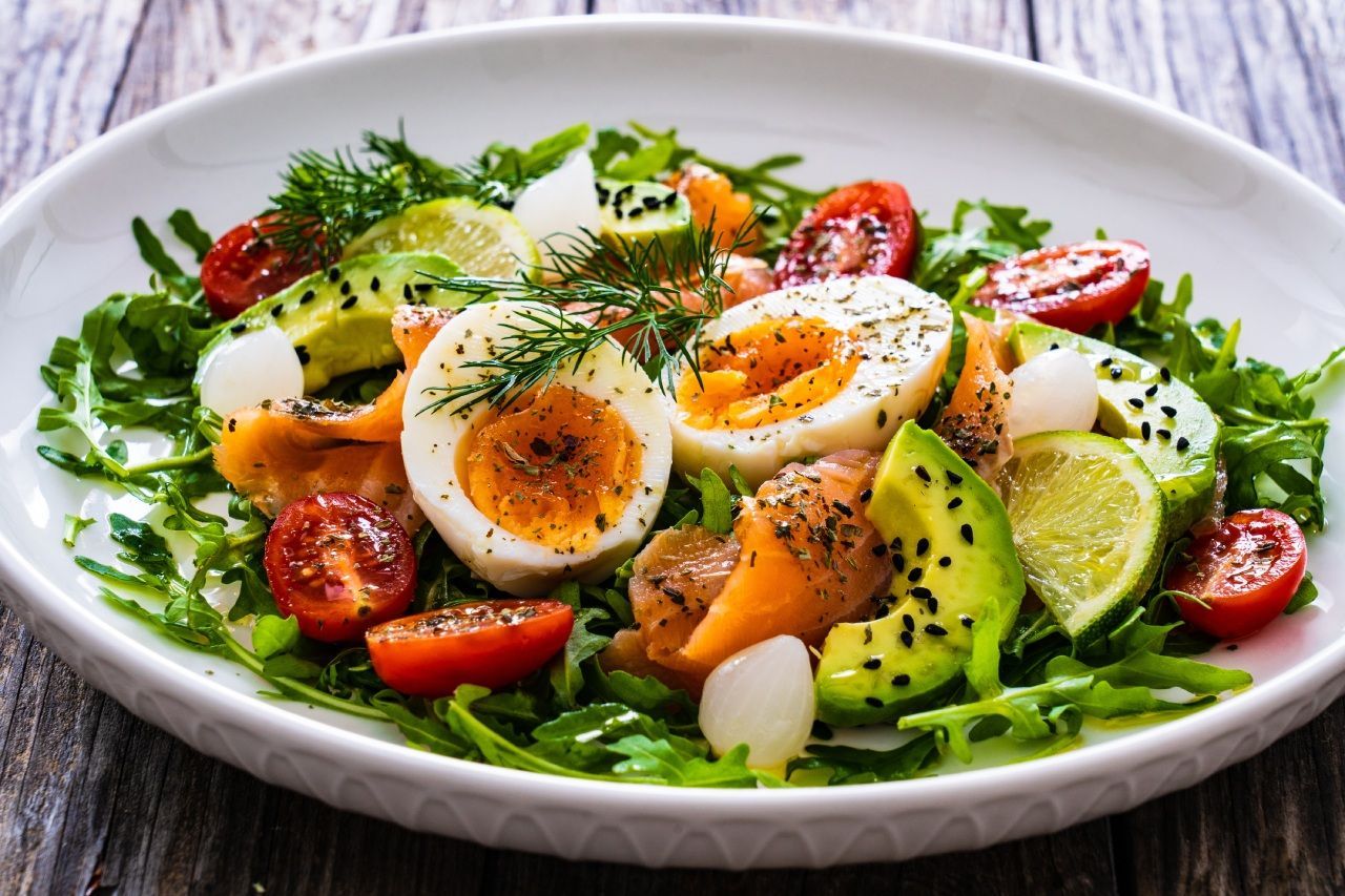 Salada colorida com folhas verdes, ovo cozido, abacate, tomate-cereja, limão e salmão defumado, temperada com ervas e sementes pretas, servida em prato branco sobre mesa rústica de madeira.