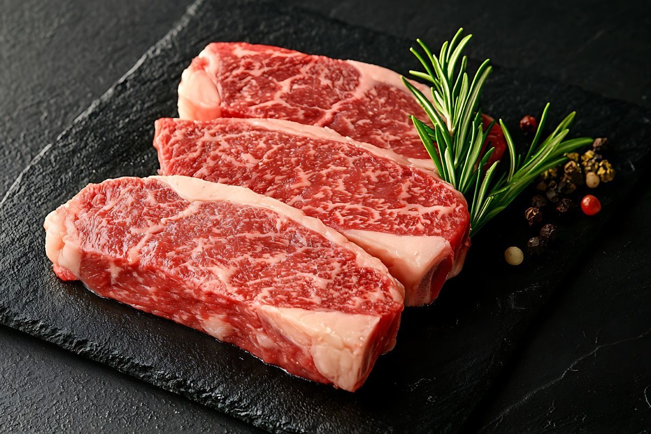 Três bifes crus em uma tábua de corte preta com alecrim e pimenta.