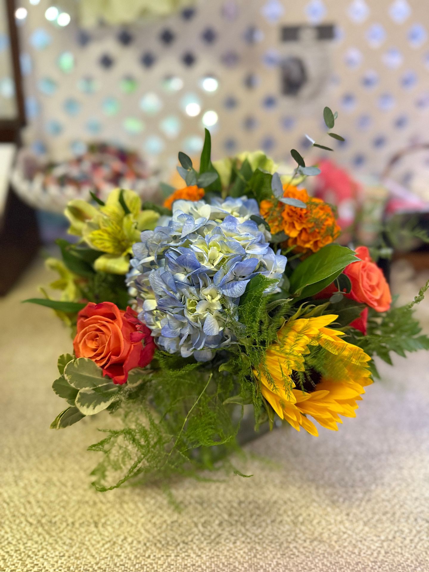 Floral arrangement with blue hydrangea, orange roses, yellow lilies, and greenery.