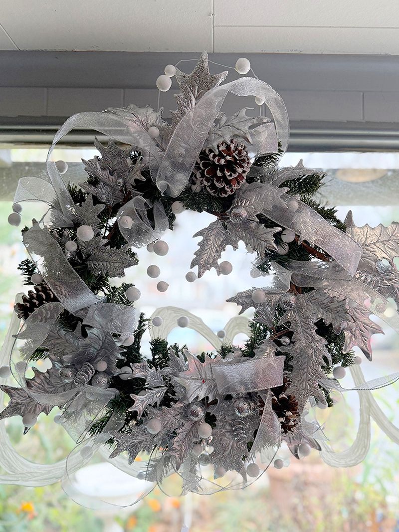 Silver and white Christmas wreath with pinecones and ribbon, hanging in front of a window.