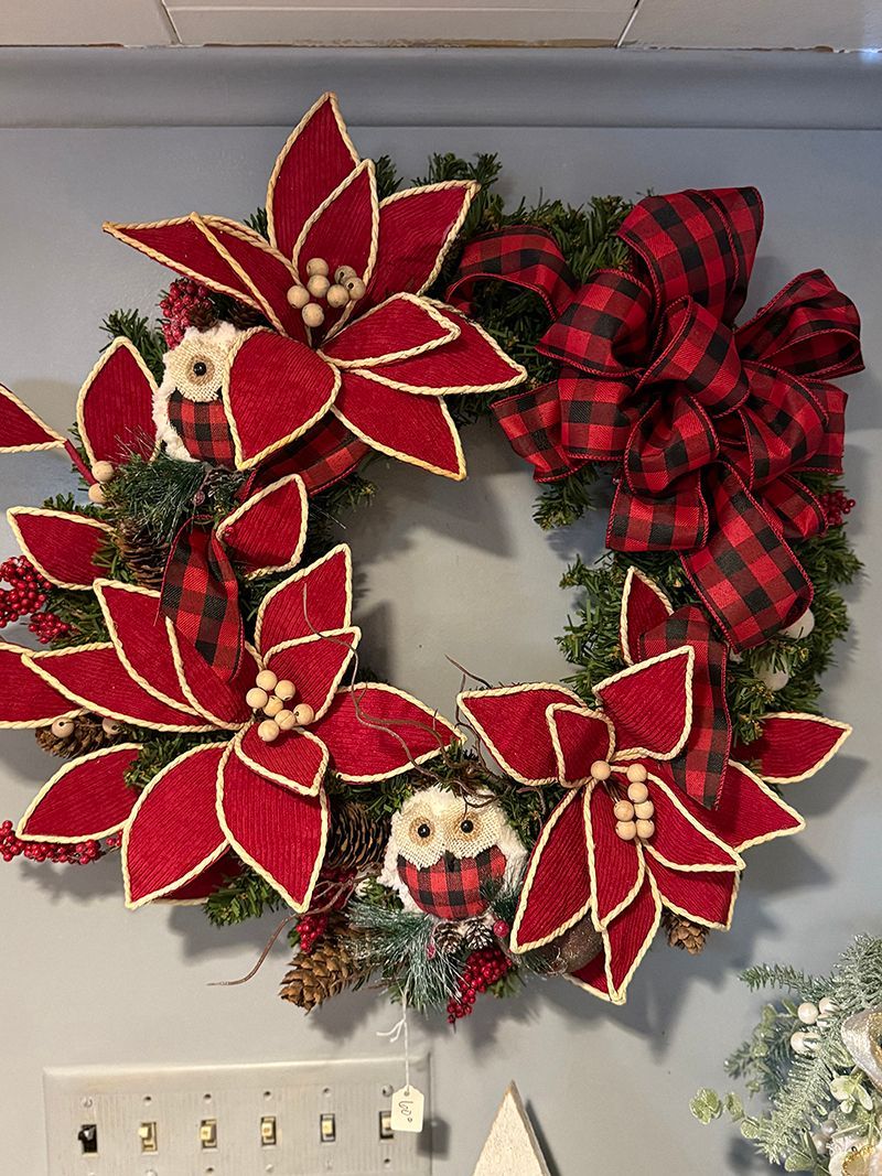Christmas wreath with red poinsettias, buffalo check bow, and owl ornaments on a wall.
