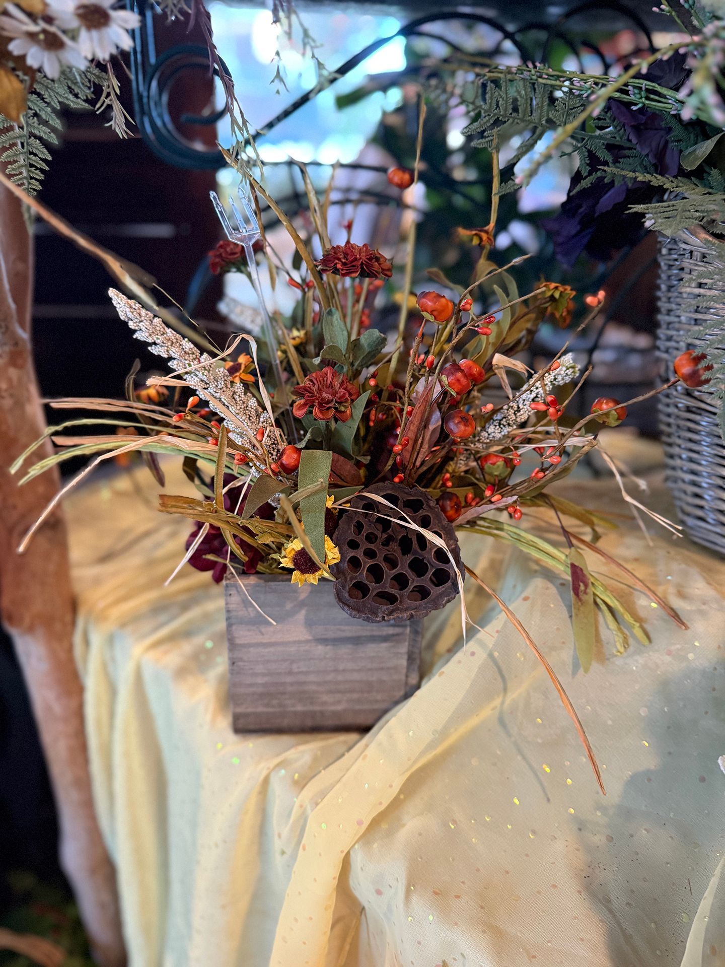 Floral arrangement in a wooden box with autumn colors: orange, brown, and green, on a yellow tablecloth.