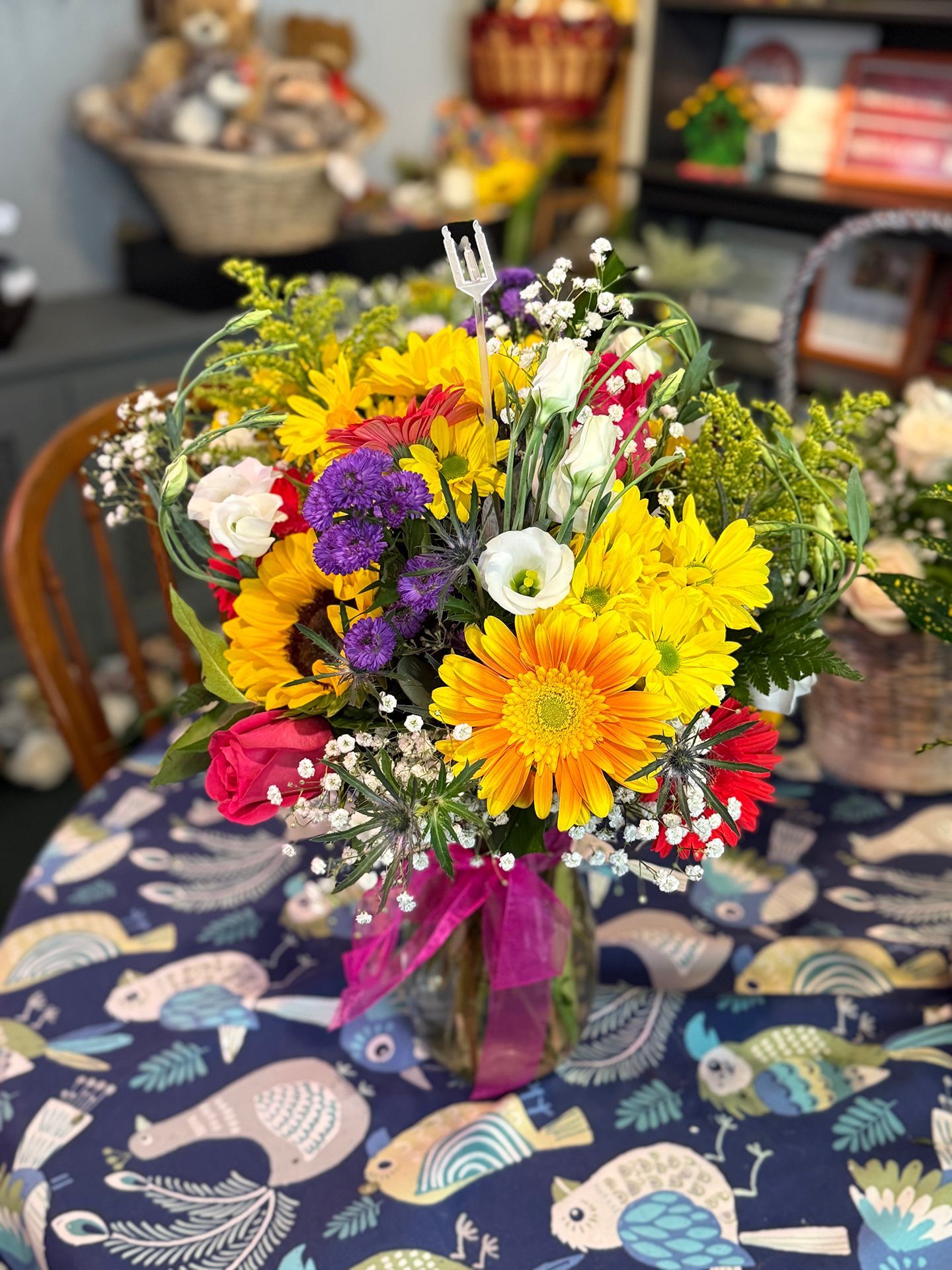 A vibrant bouquet of sunflowers, roses, and other flowers in a glass vase, tied with a pink ribbon, on a table with a bird pattern.